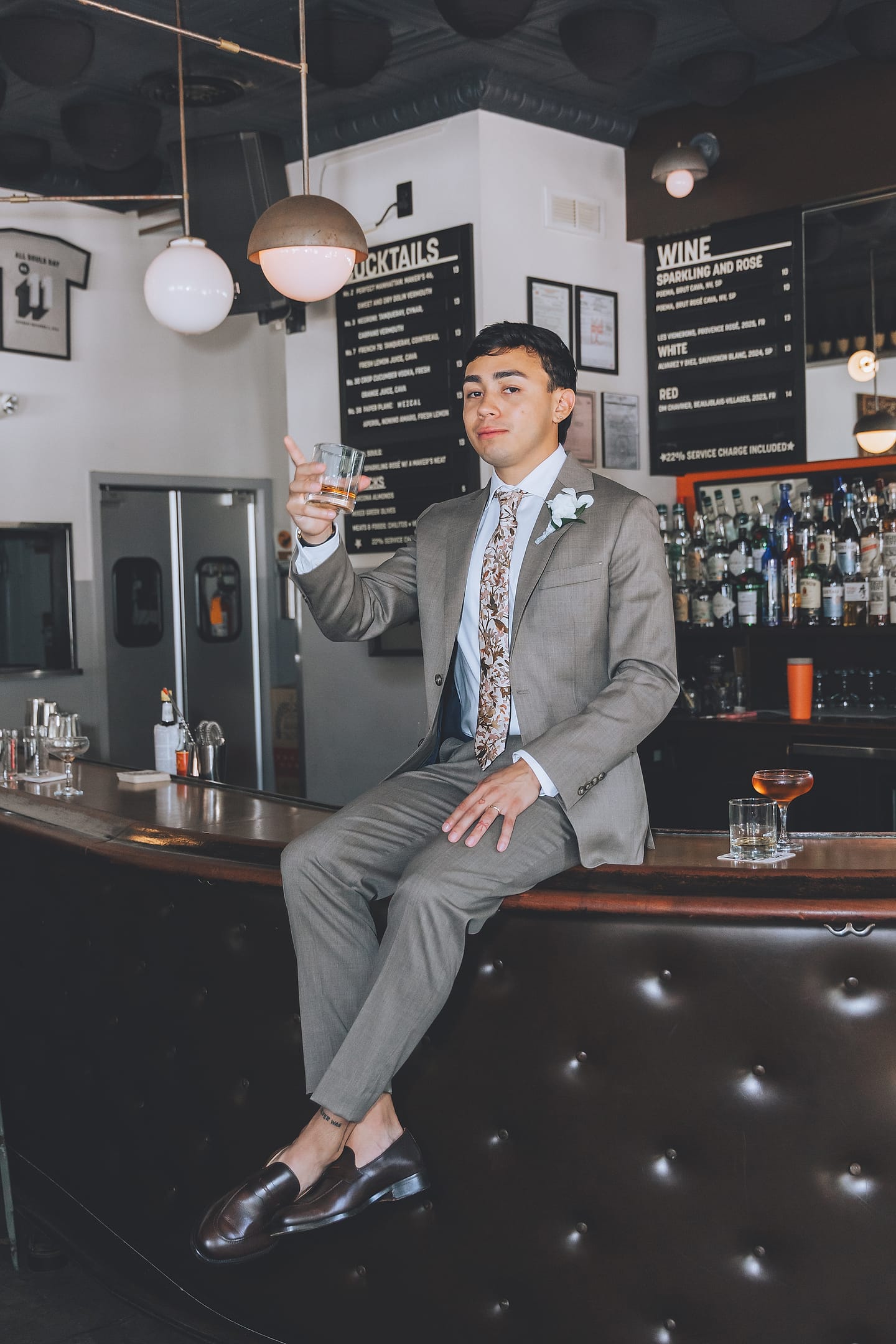 A groom raises a cocktail while seated on the bar at All Souls in Washington, D.C.