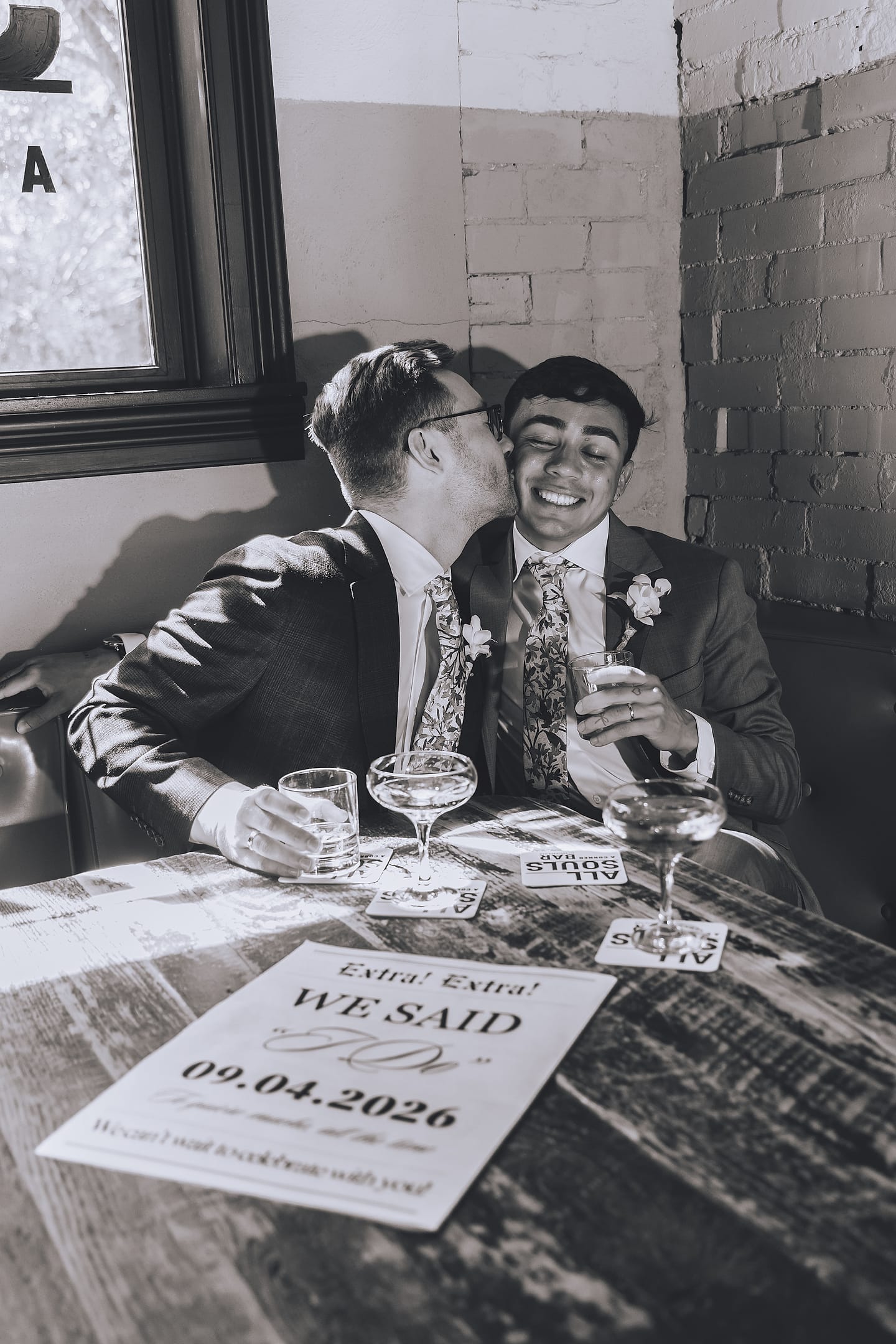 Black-and-white photo of Marco and Nick sharing a kiss over cocktails in an All Souls bar booth in Washington, D.C.