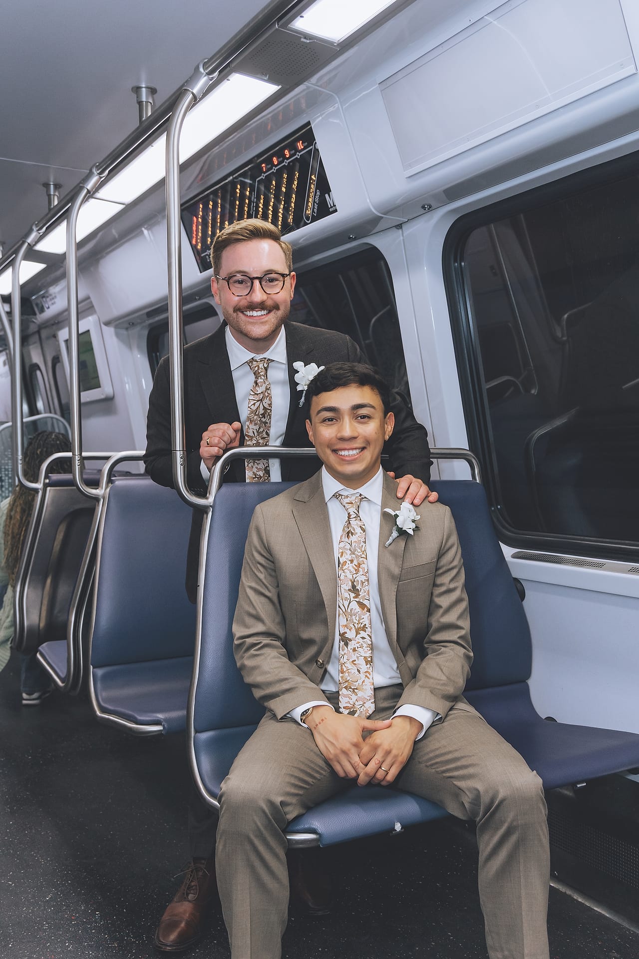 Grooms Marco and Nick in matching floral ties smile together on a Washington, D.C. Metro train on their wedding day
